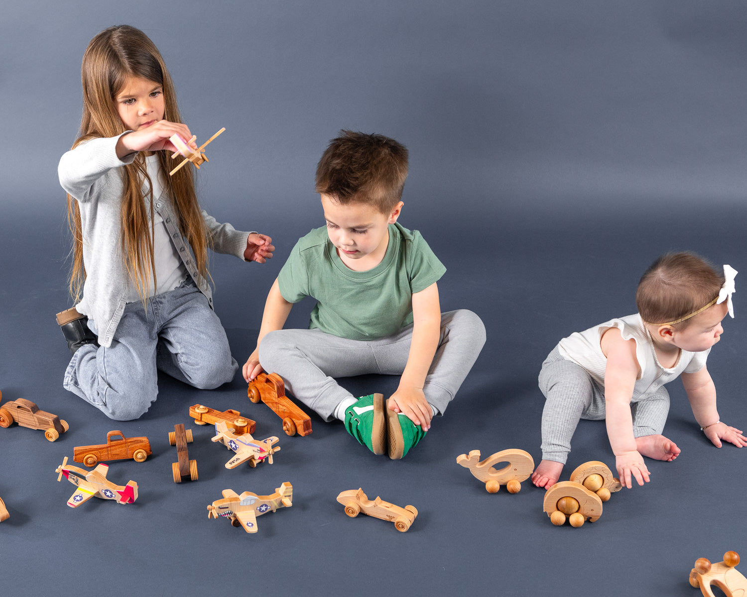 Three kids of various ages playing on the floor with wooden airplanes, cars and baby toys.