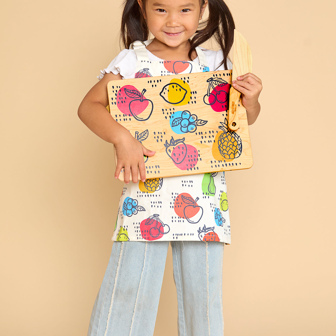 A young girl with dark hair in pigtails stands barefoot against a solid beige studio background, holding a colorful fruit-patterned cutting board with a matching fruit-print apron, and light-wash wide-leg jeans.