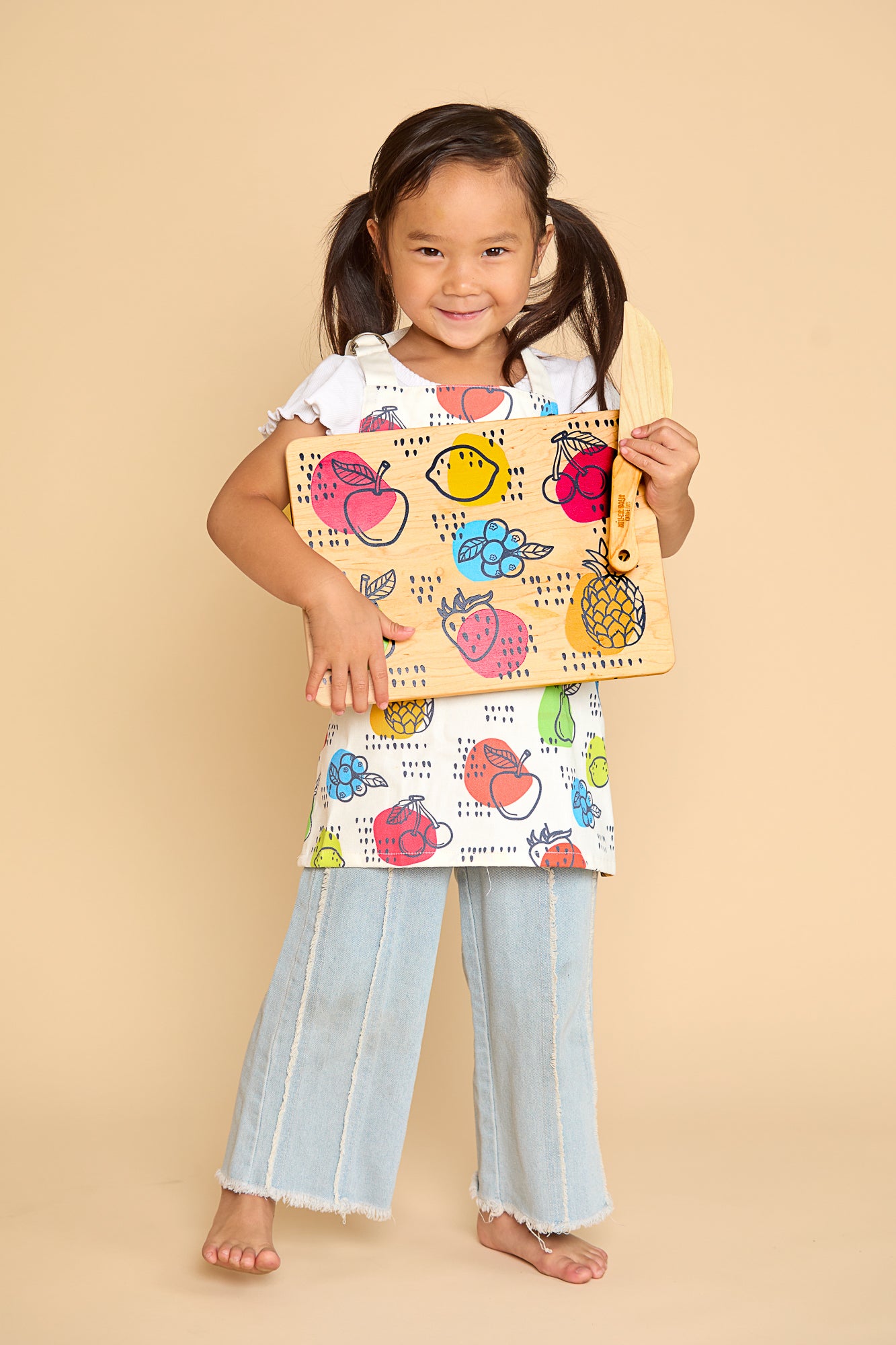 A young girl with dark hair in pigtails stands barefoot against a solid beige studio background, holding a colorful fruit-patterned cutting board with a matching fruit-print apron, and light-wash wide-leg jeans.