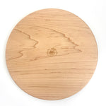 A round wooden cutting board with a natural light oak grain pattern and a small engraved logo in the center, shown on a plain white background.