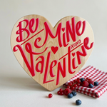 Heart-shaped wooden cutting board with red and pink script text reading 'Be Mine Valentine xoxo' on a light maple wood, styled with berries and a red-and-white checkered cloth.