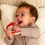 A baby lying on a white pillow, smiling and holding a red and beige heart-shaped teething toy.
