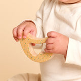 A baby wearing a cream-colored long-sleeve top holds a natural wood bird rattle and teether with green and yellow painted dots against a soft beige background.