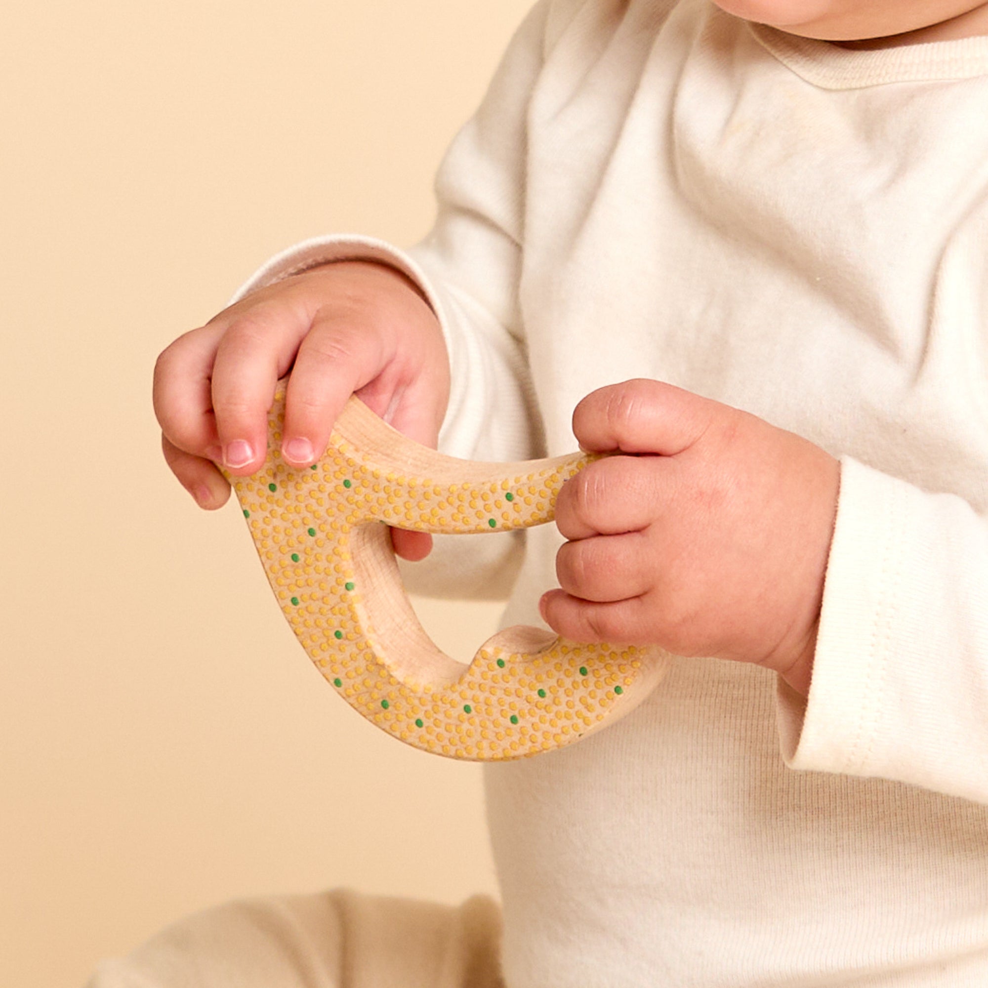 A baby wearing a cream-colored long-sleeve top holds a natural wood bird rattle and teether with green and yellow painted dots against a soft beige background.