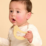 A baby wearing a cream long-sleeve top holds a yellow textured teether toy against a solid beige background.