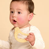 A baby wearing a cream long-sleeve top holds a yellow textured teether toy against a solid beige background.
