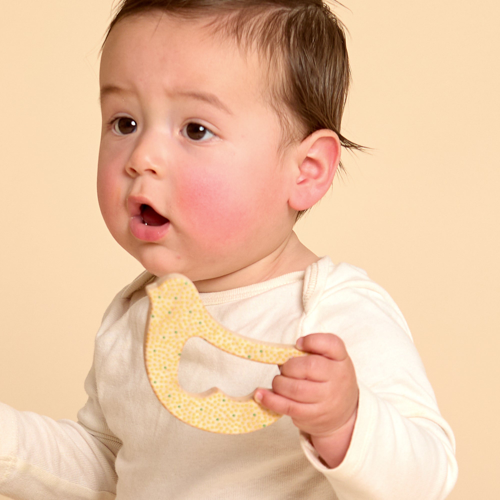 A baby wearing a cream long-sleeve top holds a yellow textured teether toy against a solid beige background.