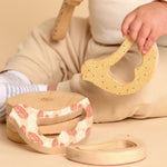 A close-up of a baby's hand holding a yellow honeycomb-patterned wooden teething toy, with two matching natural wood teething rings on a beige floor in the foreground.