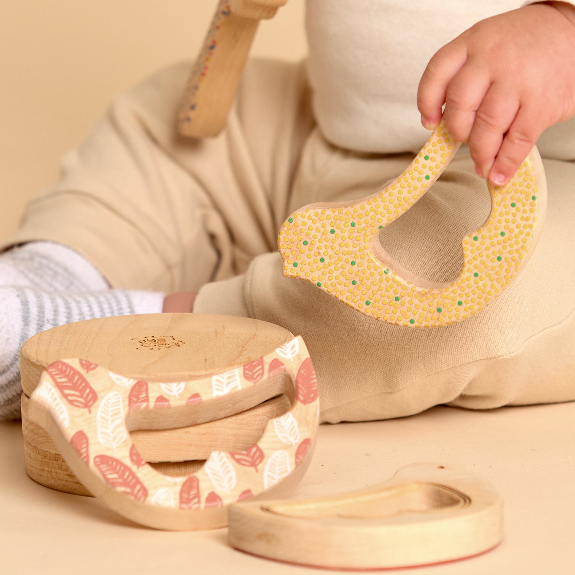 A close-up of a baby's hand holding a yellow honeycomb-patterned wooden teething toy, with two matching natural wood teething rings on a beige floor in the foreground.