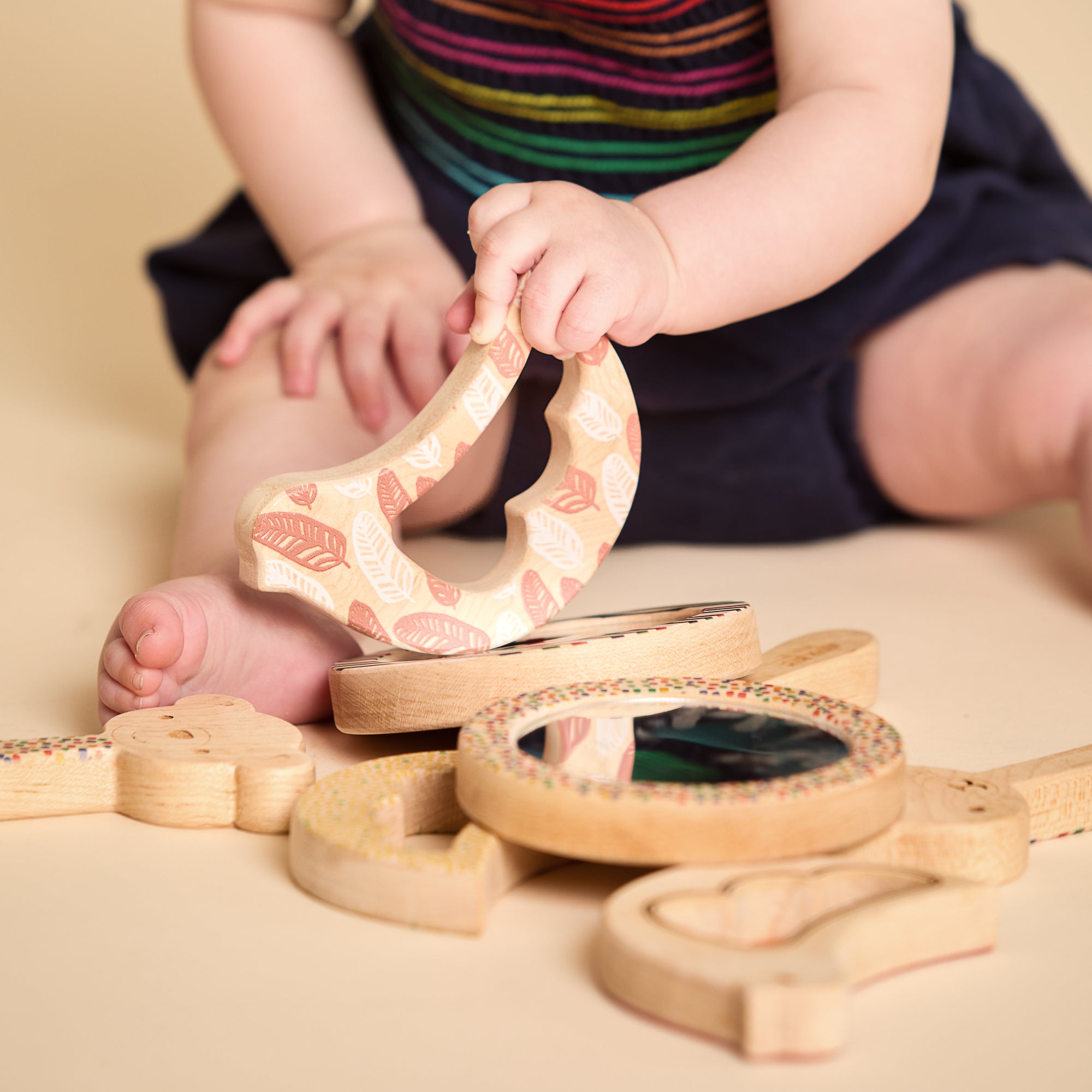 A baby sitting on a beige floor playing with a wooden sensory toy set. The baby is holding a curved wooden piece with a pink and cream leaf pattern, while other wooden toys with mirrors and colorful speckles are scattered around.