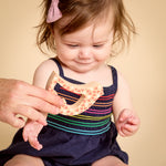 A baby wearing a navy blue dress with rainbow stripes and a pink bow in her hair, looking down at a wooden teething toy held by an adult hand against a warm beige background.