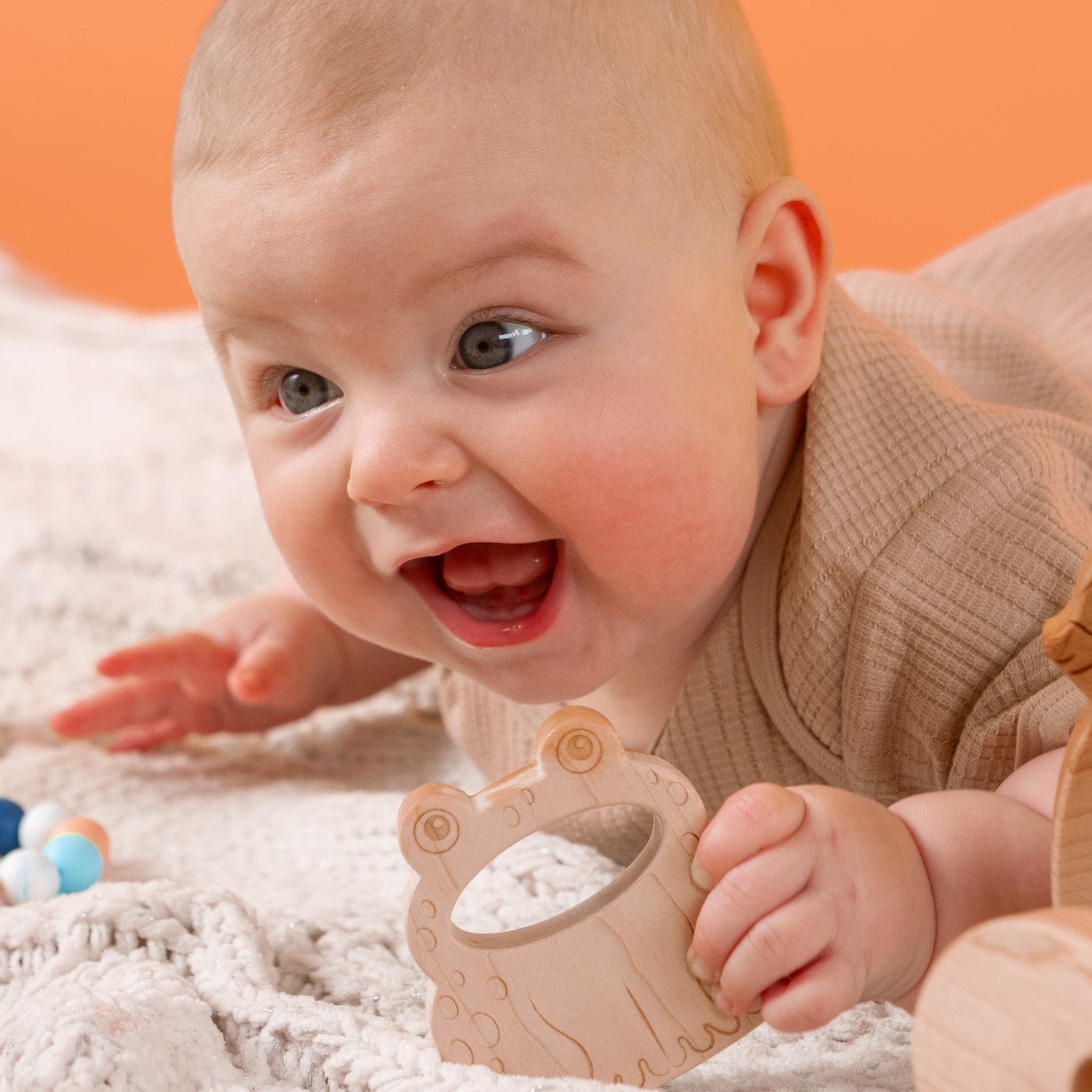 A happy baby is smiling and laughing while holding a wooden frog teether.