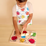 A child wearing a white apron with colorful fruit illustrations is cutting a wooden puzzle board on a light wood table against a beige background.