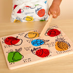 A child's hand holds a wooden toy pestle over a wooden fruit sorting board with colorful painted fruits including apples, strawberries, lemons, blueberries, pears, and pineapples.