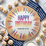 A round wooden birthday cake board with a colorful candle-style border and the words 'HAPPY BIRTHDAY' plus the name 'Emily 08-22-10', styled on a light textured surface with cupcakes, napkins, and small dishes around it.
