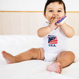 Baby wearing a onesie with 'Made in U.S.A.' text and an American flag design, sitting on a white surface.