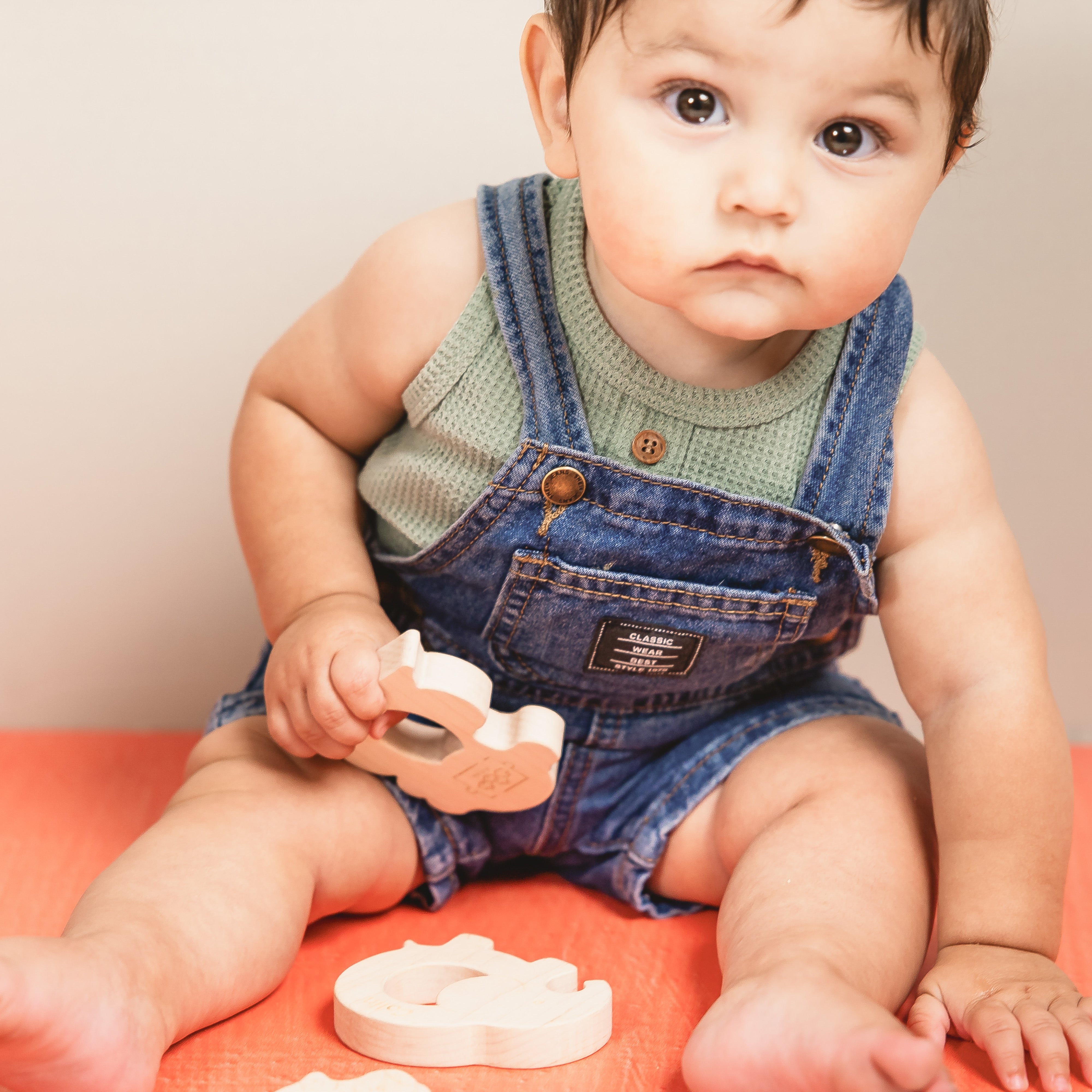Baby in denim overalls playing with wooden teethers on a red surface.