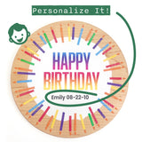 A personalized round wooden birthday board printed on light natural wood, featuring colorful painted birthday candles around the edge and the text 'HAPPY BIRTHDAY' in a rainbow gradient. A green curved line and a small green icon indicate personalization options, including the name 'Emily' and the date '08-22-10'.