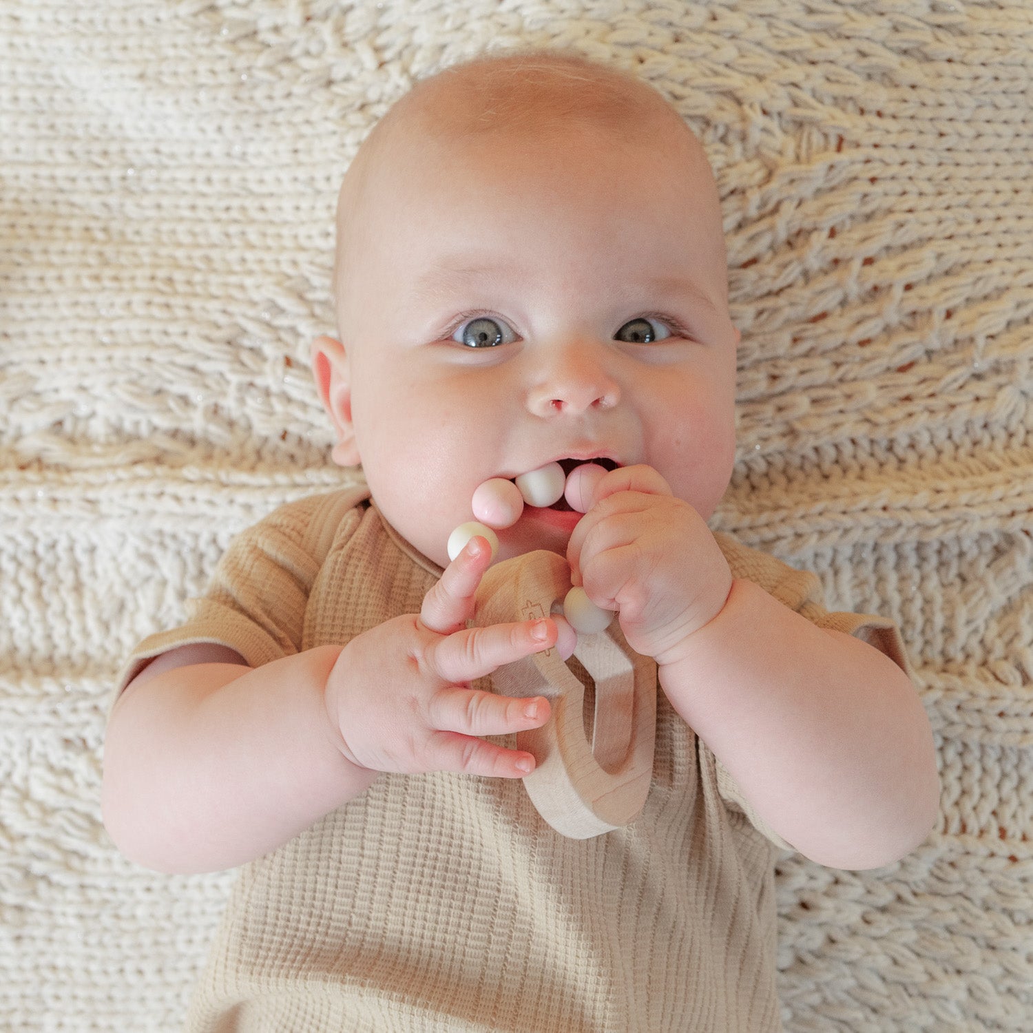 Looking from above at a baby lying on a biege blanket with a wooden heart and silicone teether.