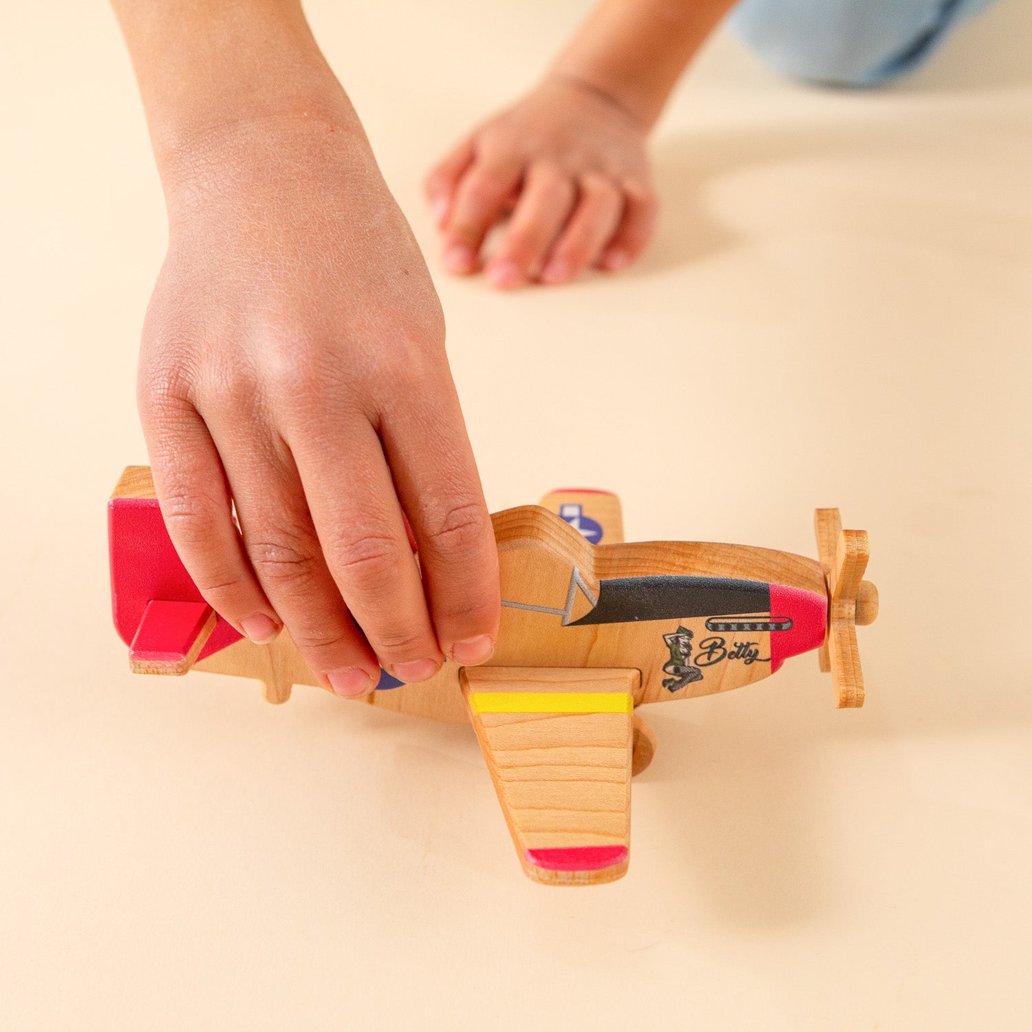 A child's hand holding a wooden P-51 Mustang airplane with a Pinup "Betty" girl near the propeller.