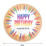 A round wooden birthday plaque with a natural wood grain border, featuring a white center with the text 'HAPPY BIRTHDAY' in a rainbow gradient and the name 'Emily 08-22-10' printed below. Colorful painted candles in a circular pattern surround the text.