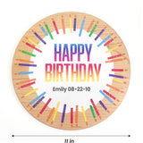A round wooden birthday plaque with a natural wood grain border, featuring a white center with the text 'HAPPY BIRTHDAY' in a rainbow gradient and the name 'Emily 08-22-10' printed below. Colorful painted candles in a circular pattern surround the text.
