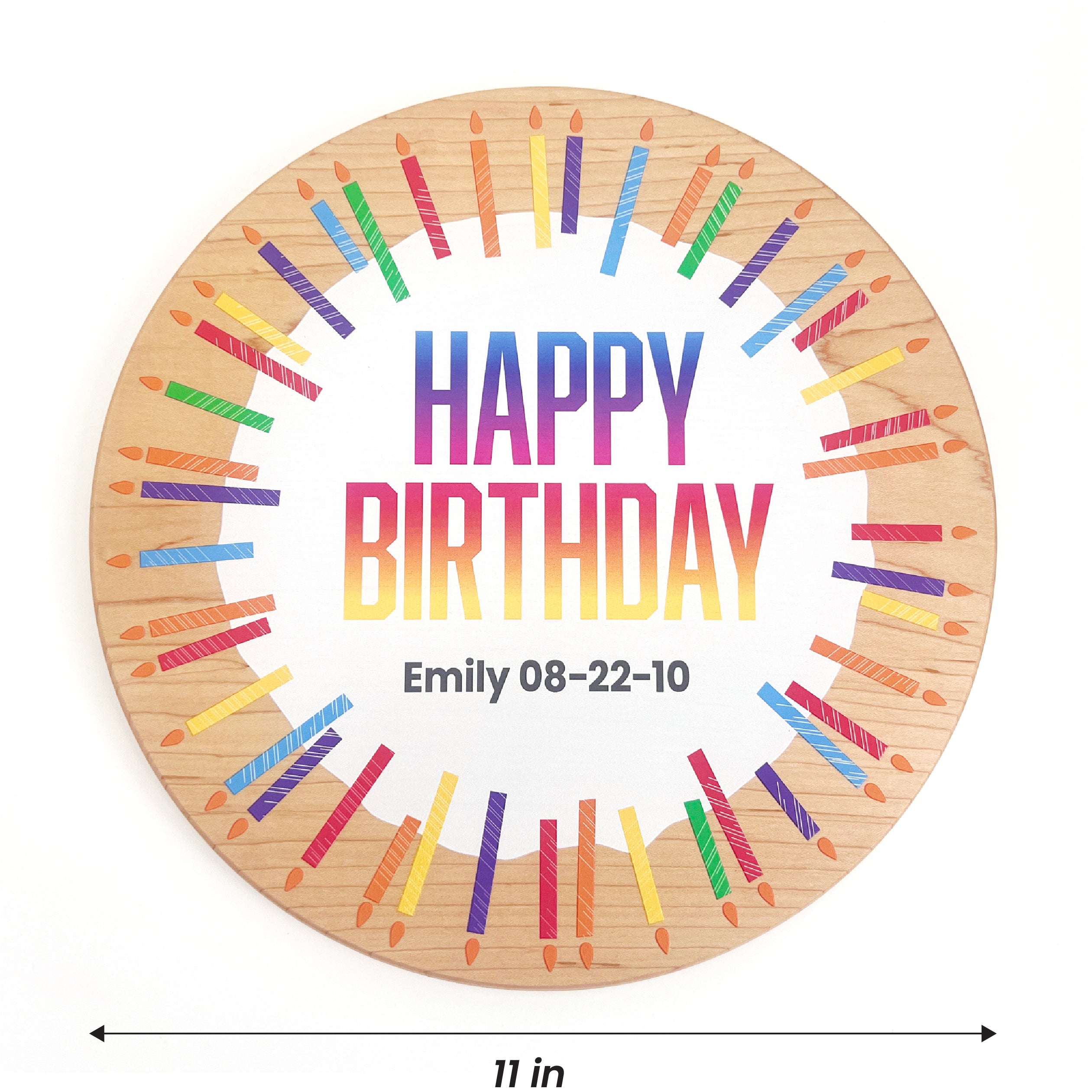 A round wooden birthday plaque with a natural wood grain border, featuring a white center with the text 'HAPPY BIRTHDAY' in a rainbow gradient and the name 'Emily 08-22-10' printed below. Colorful painted candles in a circular pattern surround the text.