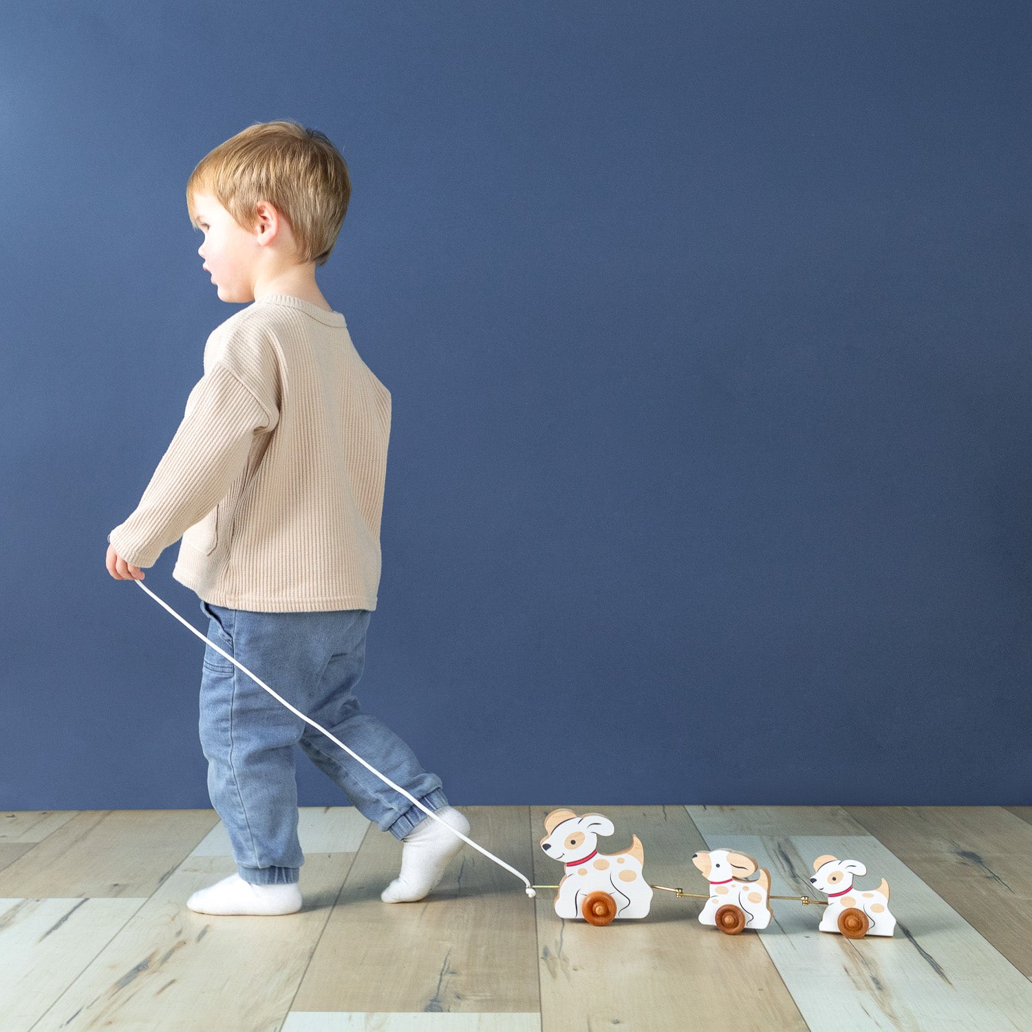 A boy holding a string pulling three wooden dogs connected on wheels behind him.