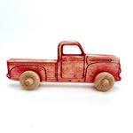 A wooden toy pickup truck with a red-painted body and natural light wood wheels, shown in profile against a plain white background.