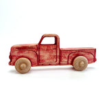 A handcrafted wooden toy pickup truck with a weathered red finish and natural light wood wheels, shown in profile against a white background.
