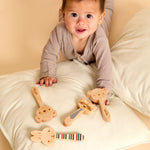 A baby in a ribbed beige long-sleeve top lies on a cream pillow, reaching toward four wooden baby teething toys with animal faces and striped accents.