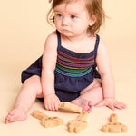 A baby girl sitting on a beige studio floor wearing a navy blue romper with colorful embroidered stripes across the chest, playing with wooden toy pieces.