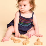 A baby girl sitting on a beige studio floor wearing a navy blue romper with colorful embroidered stripes across the chest, playing with wooden toy pieces.