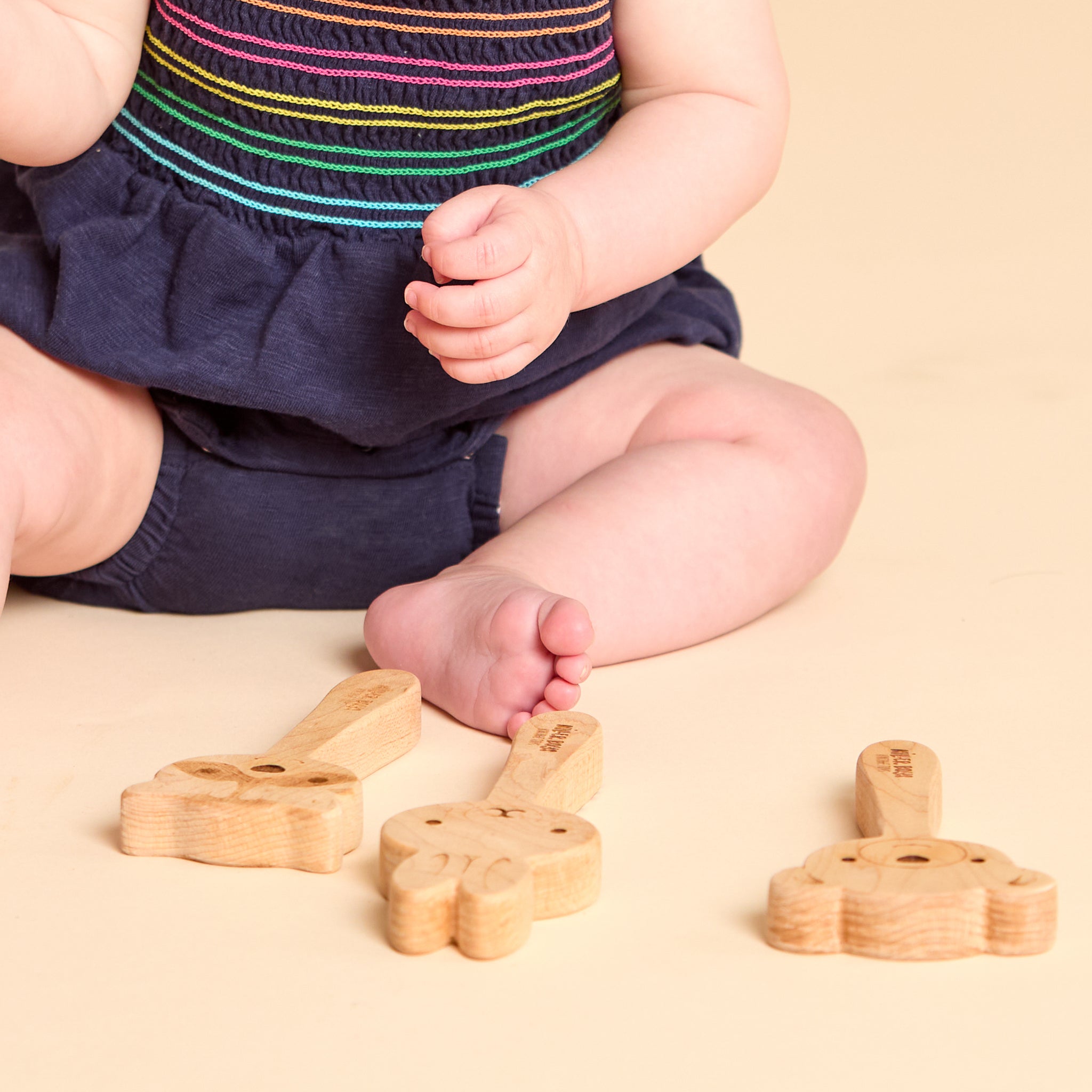 A baby sitting on a beige floor wearing a navy blue romper with colorful horizontal stripes. In front of the baby are three small, light natural wood animal-shaped toys, including a rabbit and a duck.