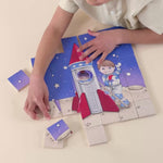 A boy laying on the ground putting together a wooden magnetic piece puzzle of an astronaut in space.
