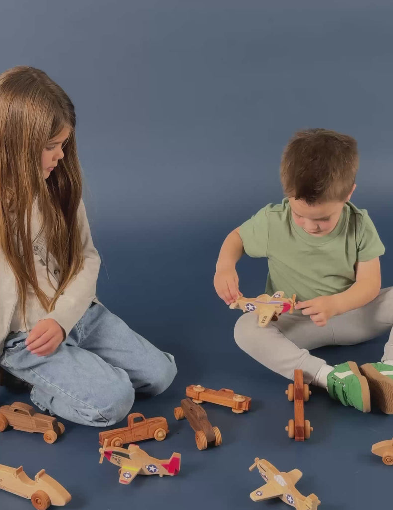 A brother and sister sitting on the floor playing with P-51 Mustang wooden airplanes and wooden trucks.