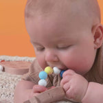 A baby lying on a blanket teething on a yellow white and blue silicone and wooden teether.