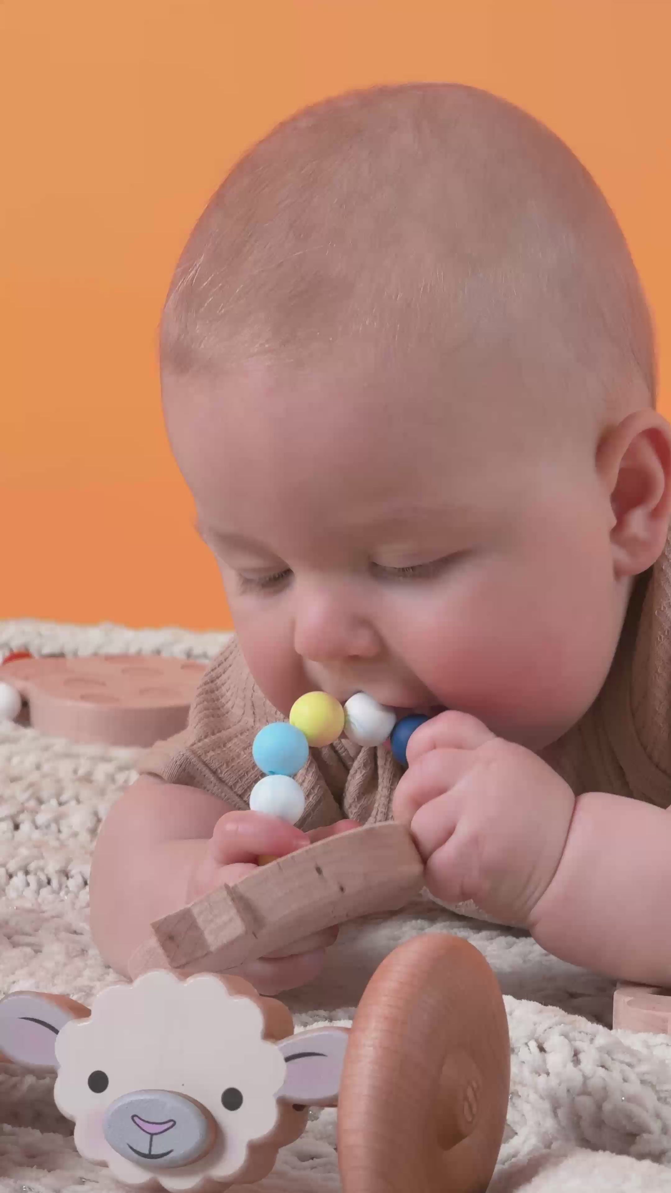 A baby lying on a blanket teething on a yellow white and blue silicone and wooden teether.